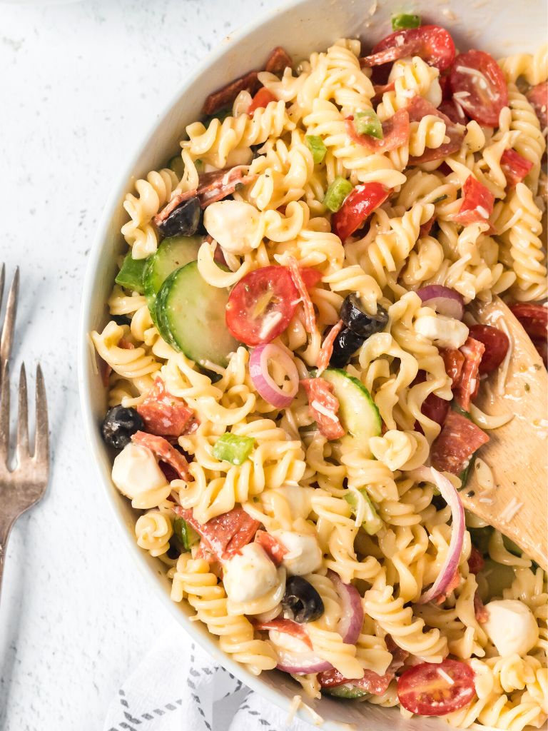 White bowl with pasta salad inside of it and a wooden spoon on the side.