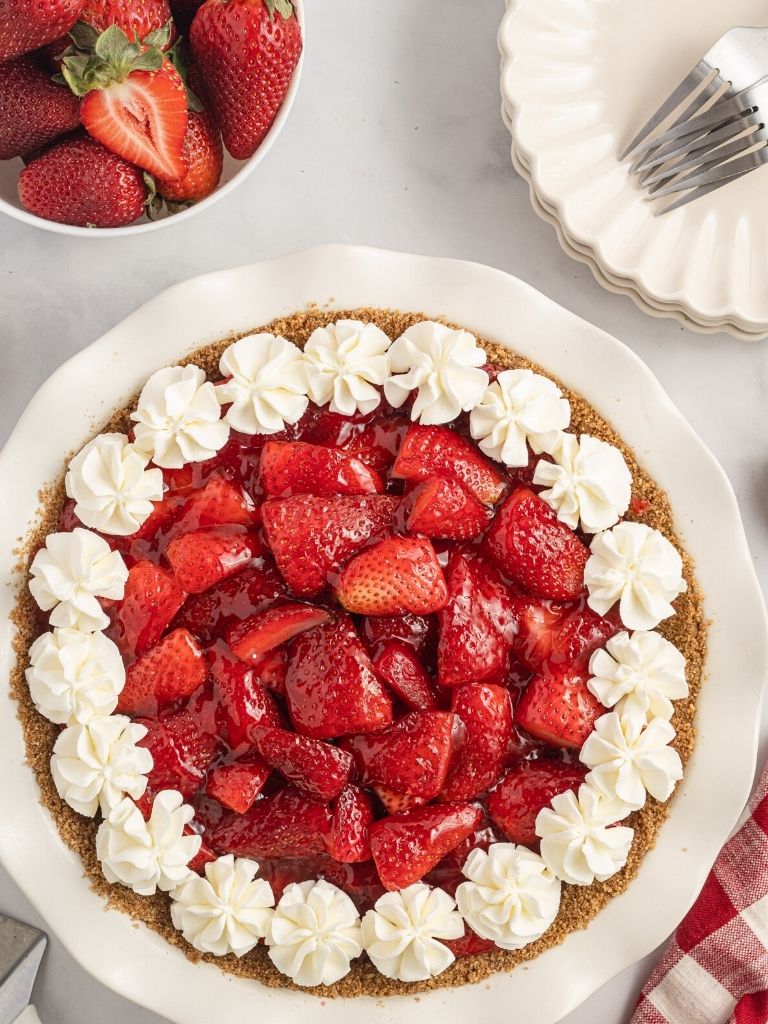 Overhead picture of a pie topped with jello and whipped cream. Stack of plates on the side and a bowl of strawberries.