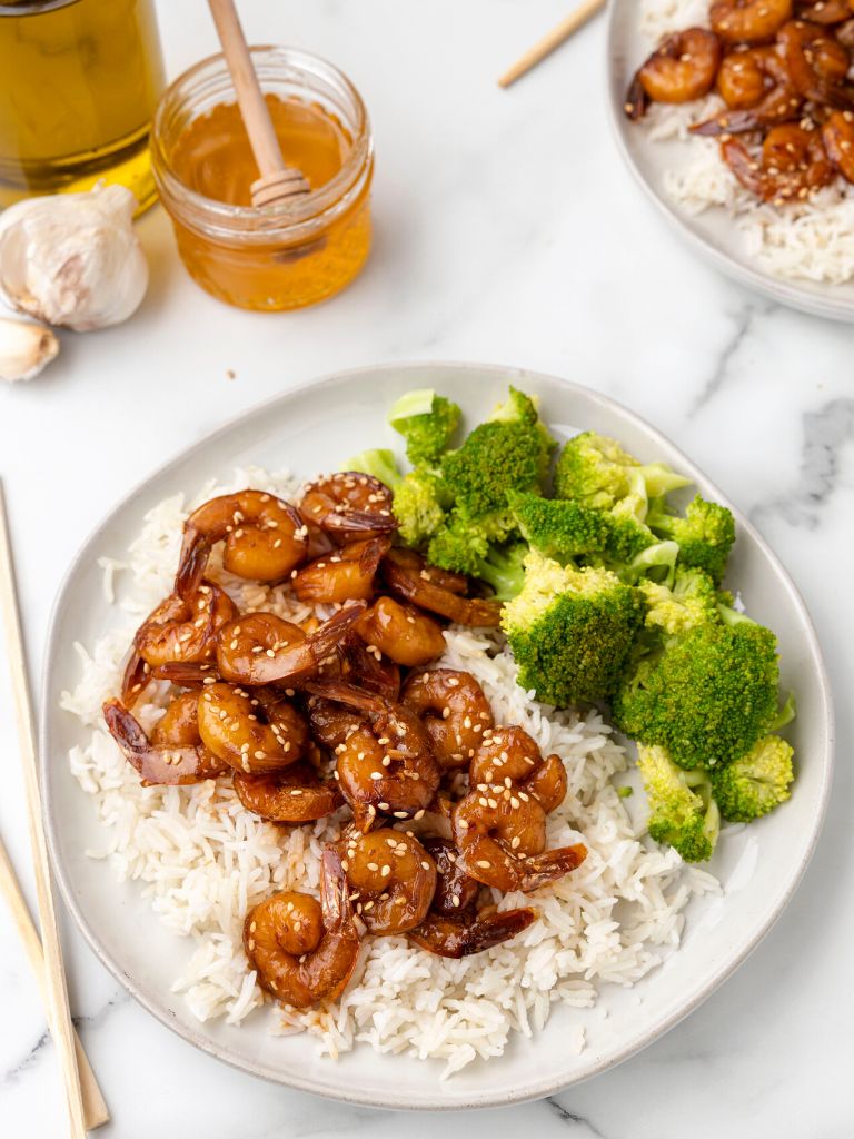 Overhead view of a serving of rice topped with shrimp and broccoli on the side. Chopsticks and a jar of honey to the side of it. 