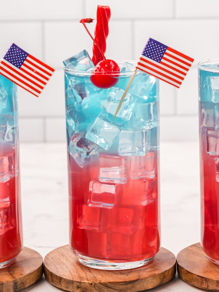 Layered drink for 4th of July with a licorice rope inside of it, topped with a cherry and an American flag toothpick. 