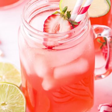 A clear glass of strawberry limeade garnished with lime slices, strawberries, and two paper straws.