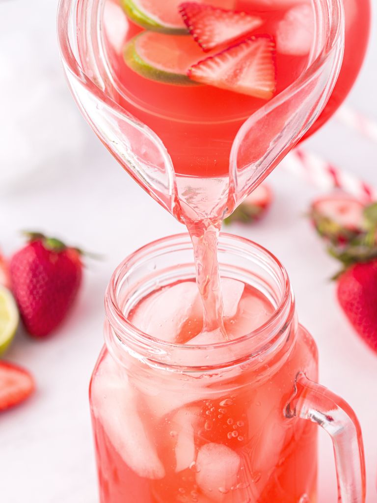 A cup of strawberry limeade with a pitcher above it pouring punch unto the cup. 