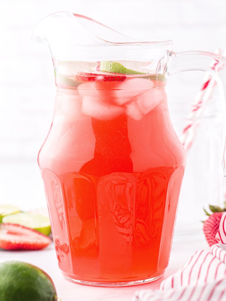 A glass pitcher of limeade with strawberries and lime slices on top.