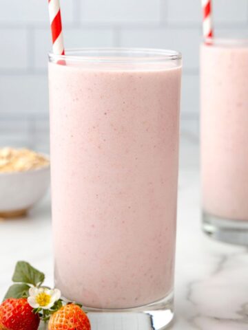 A clear glass with strawberry oatmeal smoothie inside of it, a red and white straw, and strawberries at the base of the glass cup.