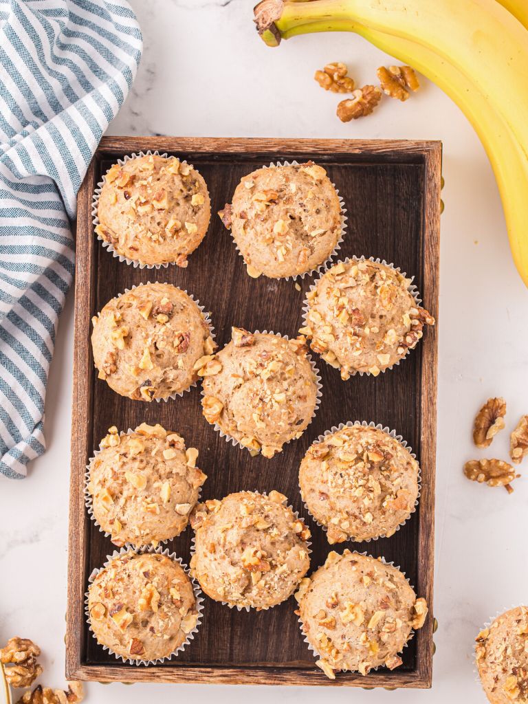 Overhead shot of muffins on a wooden tray with a banana and chopped nuts to the side, along with a blue striped cloth. 