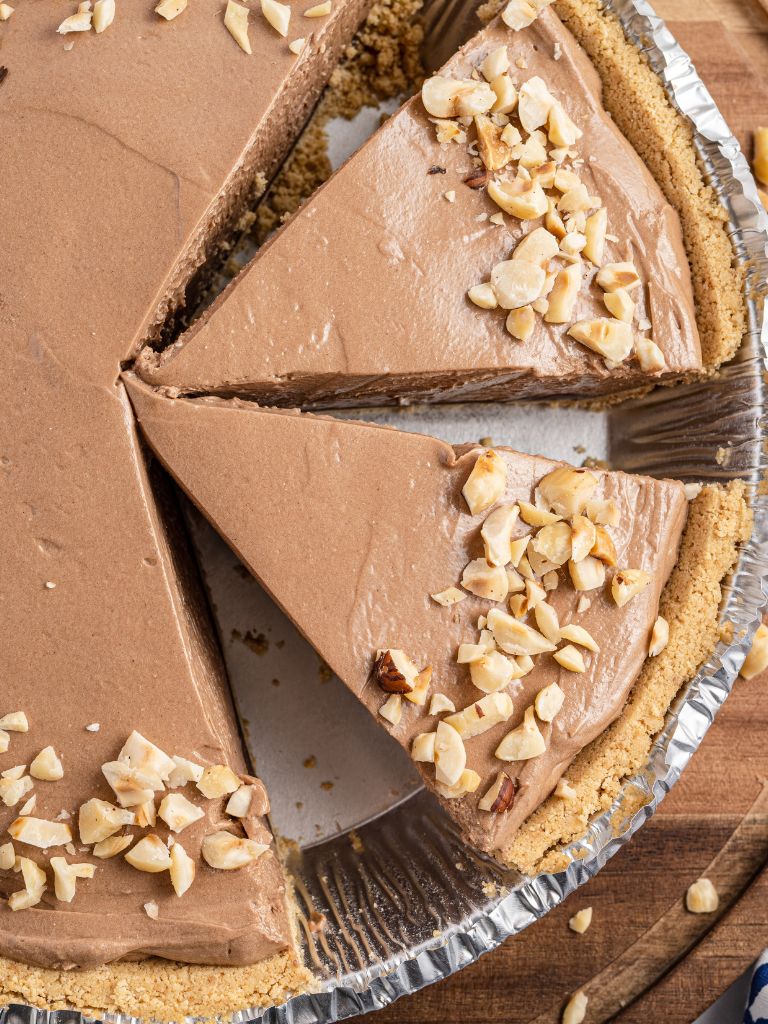 Overhead shot of a pie topped with hazelnuts, cut into slices on a wooden background.