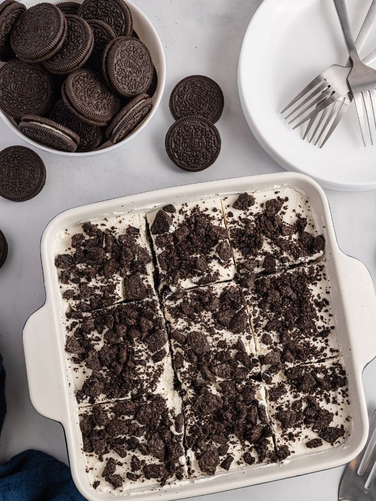 Overhead shot of a pan of icebox cake with oreo cookies on the side, a stack of plates and forks for serving. 