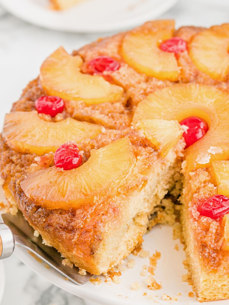 A slice of cake with pineapple and cherries on top, a spatula underneath it pulling it from the cake pan. 