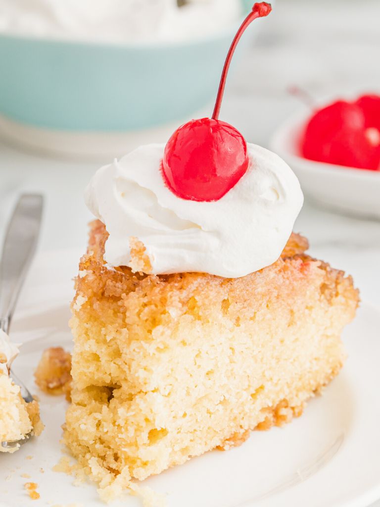 A slice of pineapple upside down cake topped with whipped cream and a cherry. Slice of cake is sitting on a white plate with a fork to the side. 