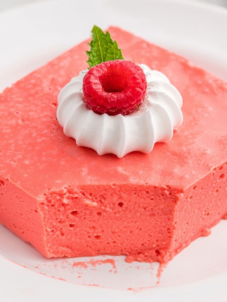 A square of jello salad on a white plate topped with whipped cream and a fresh raspberry. 