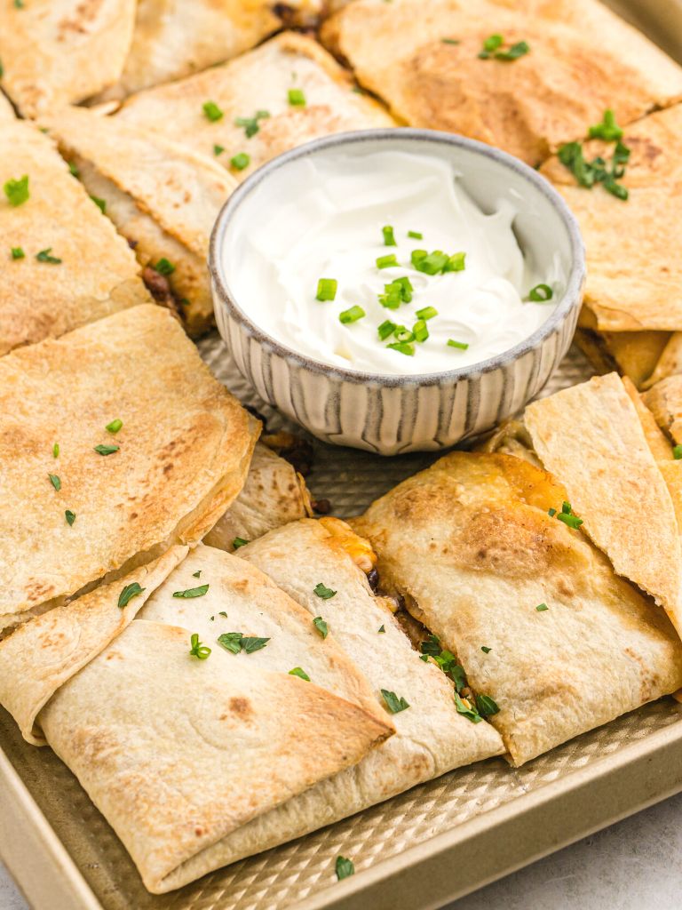 A sheet pan of flour tortilla quesadillas with a bowl of sour cream in one spot. 