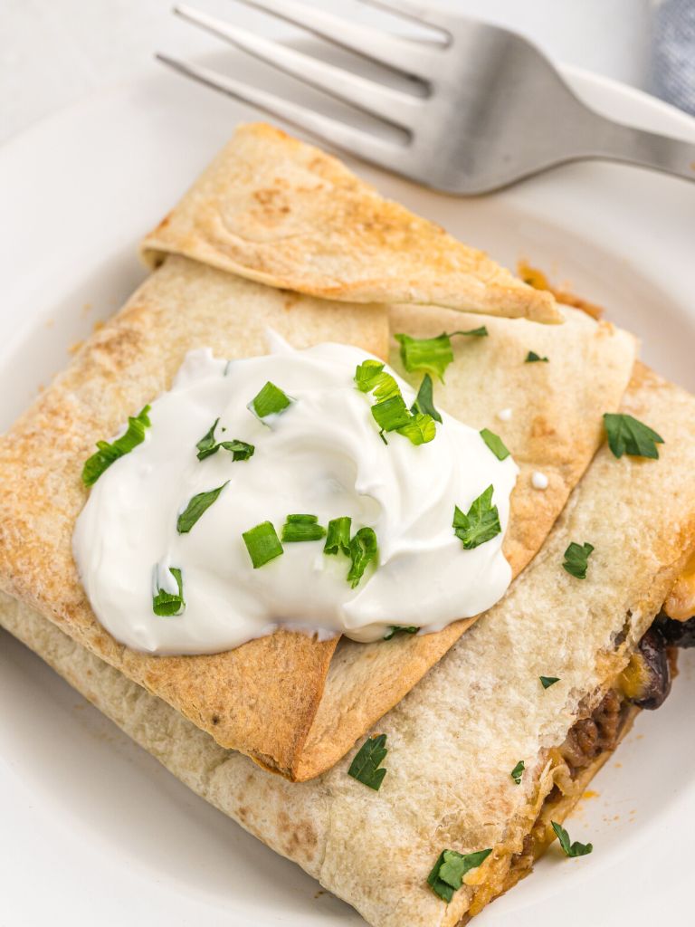 One square of a crispy quesadilla on a white plate topped with sour cream and cilantro. 