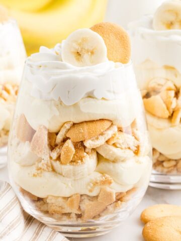 A glass cup with layers of banana pudding, whipped cream, bananas, and Nilla Wafers. Topped with a banana and Nilla Wafer on top for garnish.