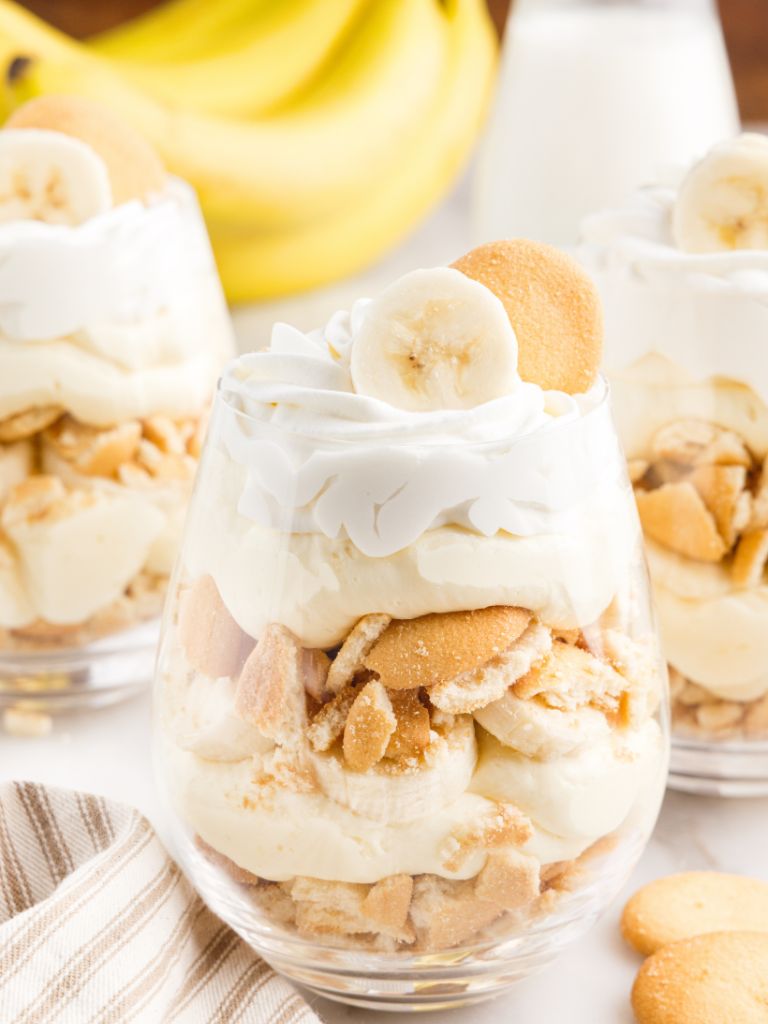 Glass cups with banana pudding parfait layers inside, with bananas and a glass of milk in the background.