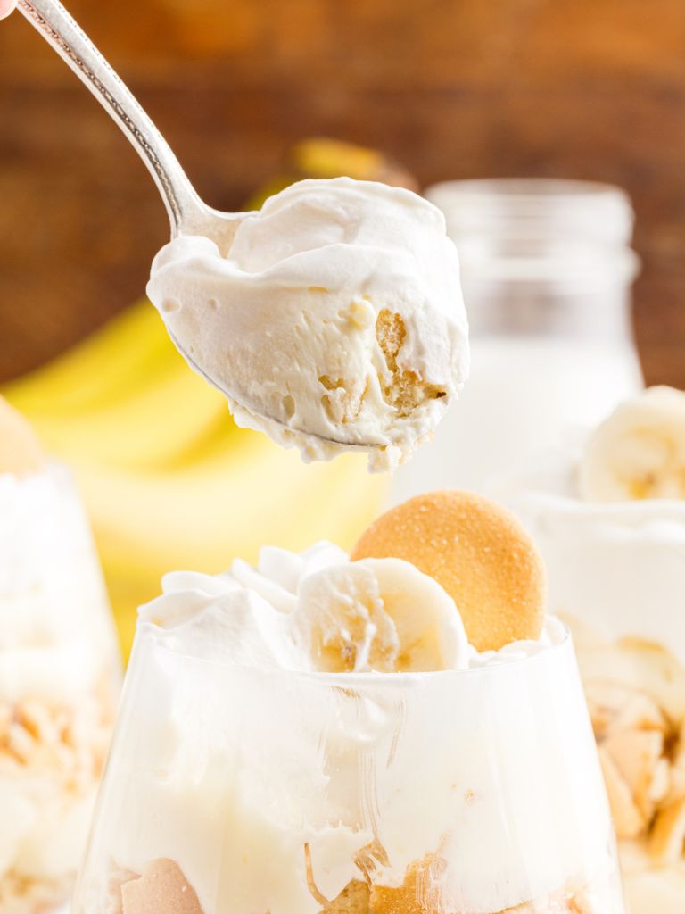 A bite of banana pudding and whipped cream on a spoon, above a glass cup of parfait inside of it. 
