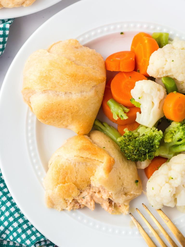 A dinner plate with vegetables, a fork, and a crescent chicken pillow on it. 