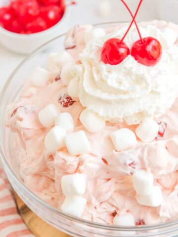 A glass bowl with fluff and cherries inside of it and topped with whipped cream and cherries on top.