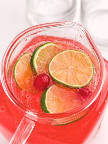 A glass pitcher filled with cherry limeade drink with lime slices and a cherry on top of the drink.