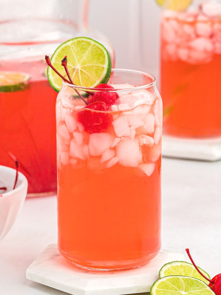 A tall glass with pink punch inside of it, topped with a lime slice and a cherry. A green straw in the background.
