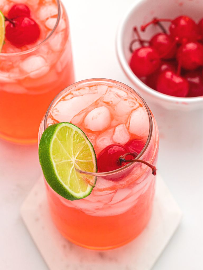 A cup with limeade inside of it with cherries and a lime. 