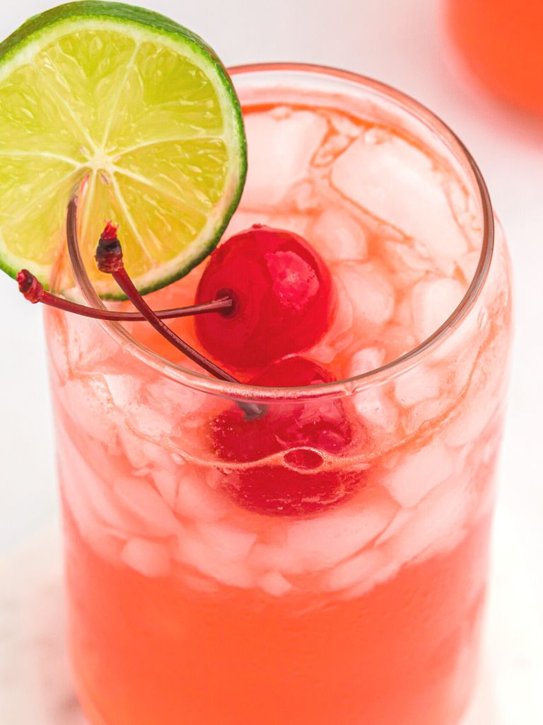 A close up shot of a drink with lime and a cherry on top, inside a glass cup. 