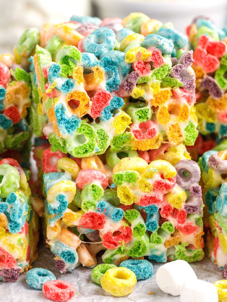 A stack of two cereal bars with Froot Loops cereal on top of each other, some marshmallows and cereal to the side of the stack.