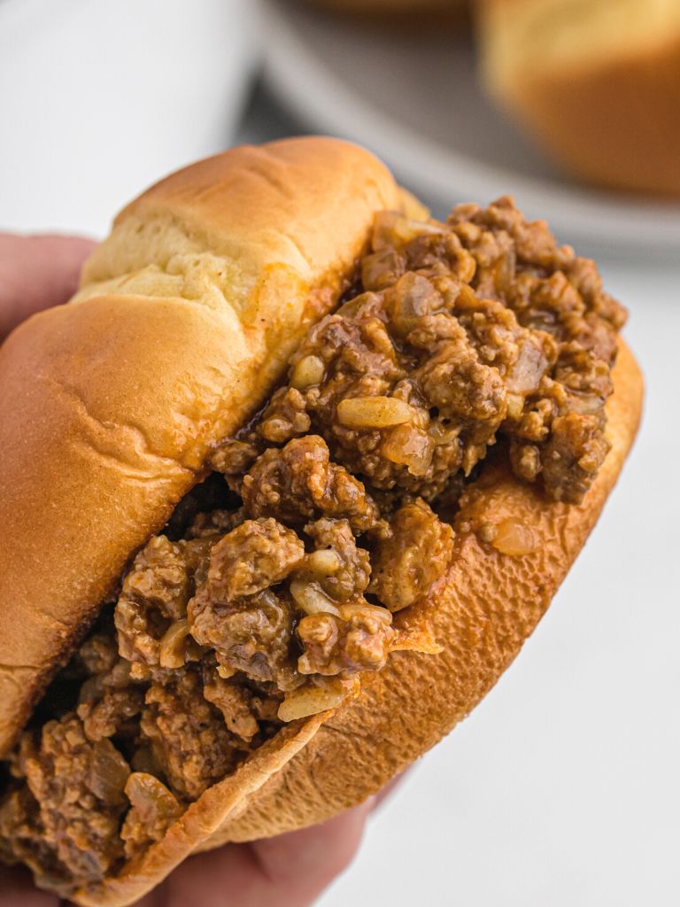 A sloppy Joe sandwich being held in a hand, sideways, so the viewer can see the filling inside of it.