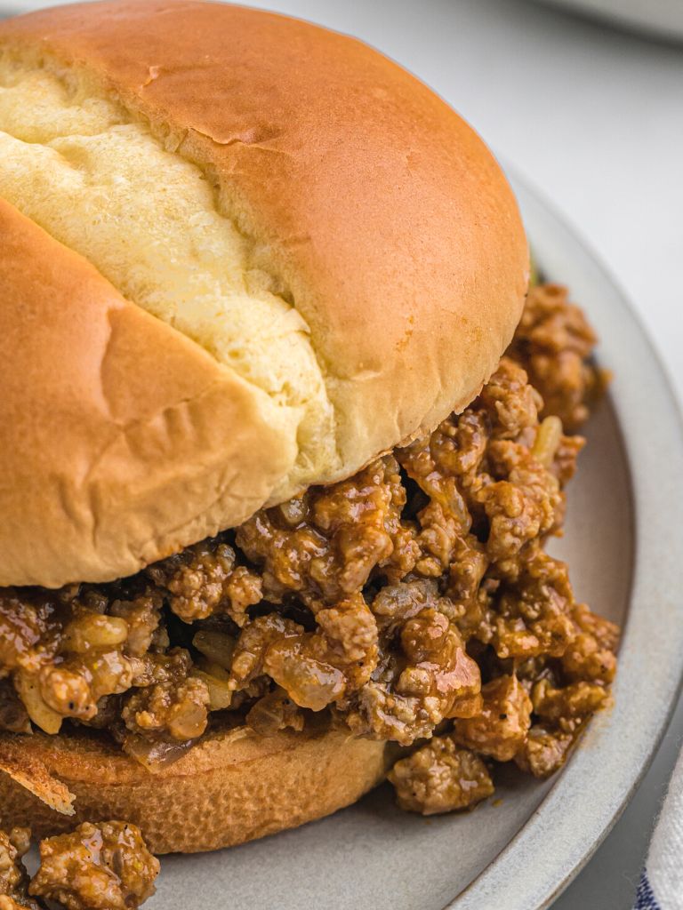 A hamburger bun filled with sloppy Joe meat mixture spilling out the sides of it. On a gray dinner plate.