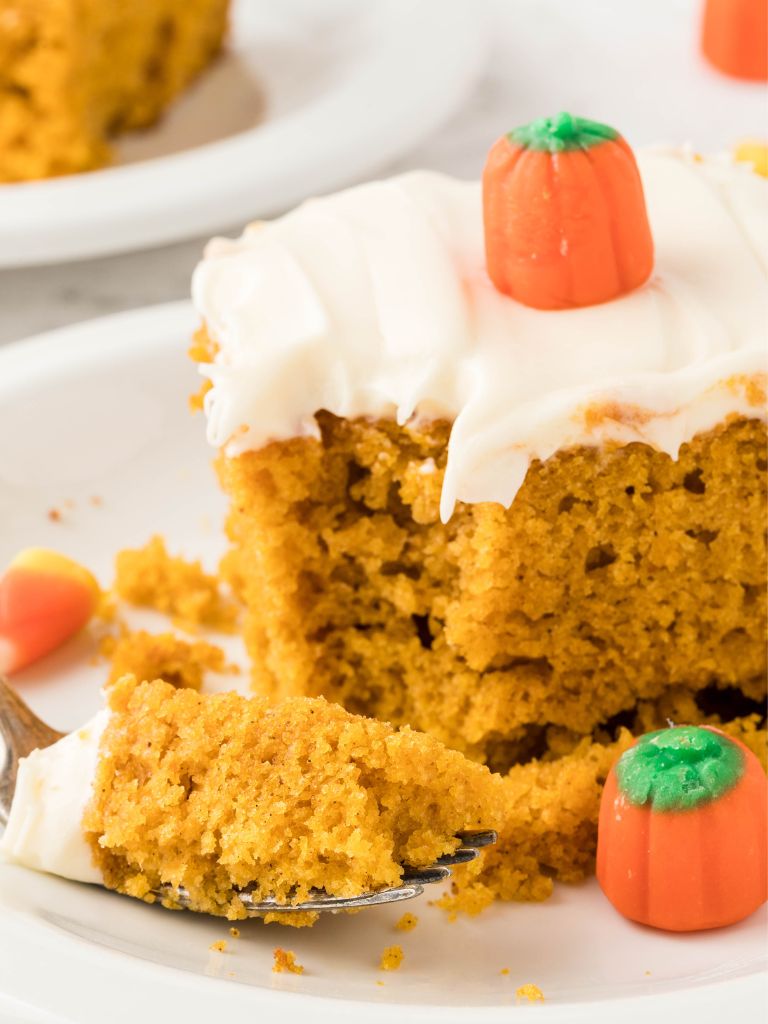 A piece of cake with frosting and a marshmallow pumpkin on top, a fork with a bite on it also on the white plate. 