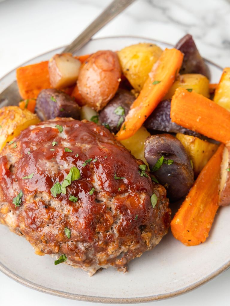 A plate with meatloaf and roasted vegetables on it, with a silver fork on the side. 