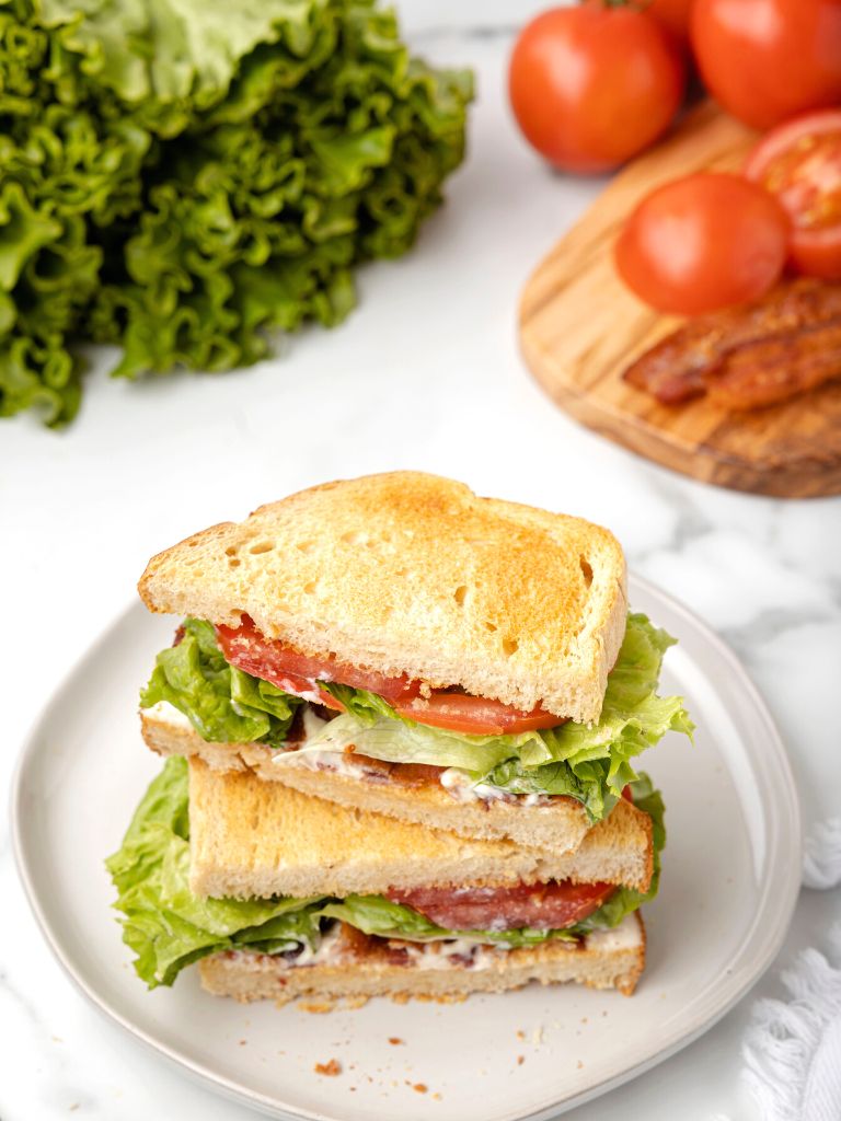 A blt sandwich on a white plate with tomatoes and lettuce to the side of the plate.