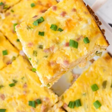 A white spatula holding a piece of breakfast casserole with eggs, ham, and potatoes.