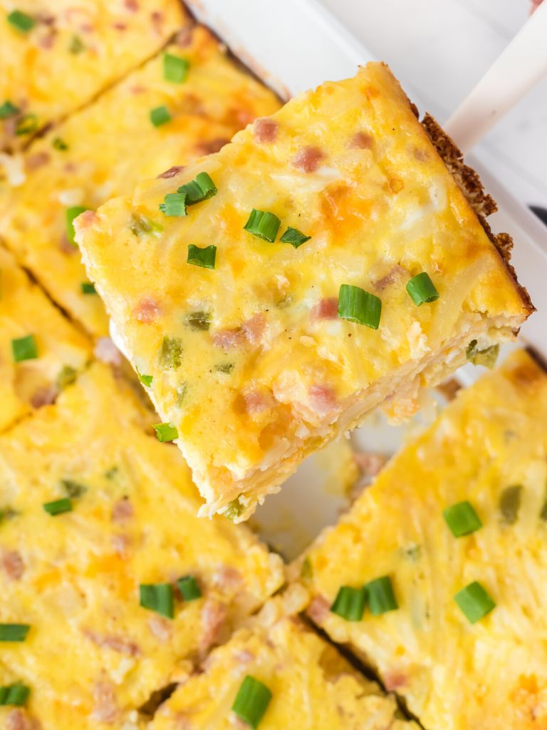 A white spatula holding a piece of breakfast casserole with eggs, ham, and potatoes. 