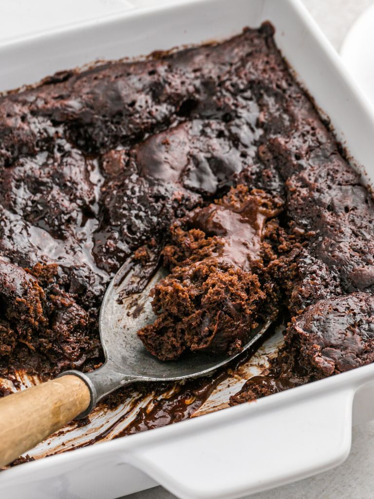 A white baking dish of chocolate cake with fudge sauce and a spoon inside the cake.