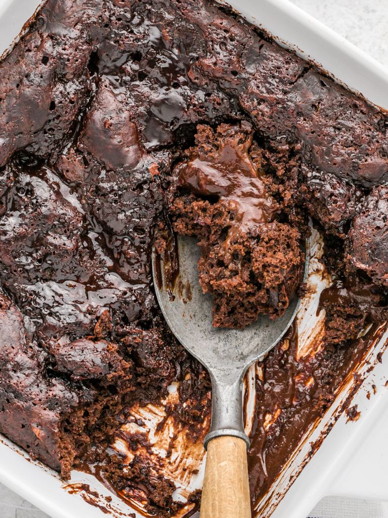 A baking dish of chocolate cake with a serving spoon inside taking a scoop.
