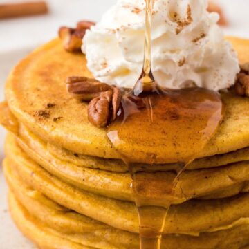 A stack of pancakes on a white plate topped with maple syrup drizzling down the sides, pecans, and whipped cream.