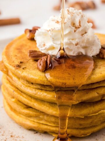 A stack of pancakes on a white plate topped with maple syrup drizzling down the sides, pecans, and whipped cream.