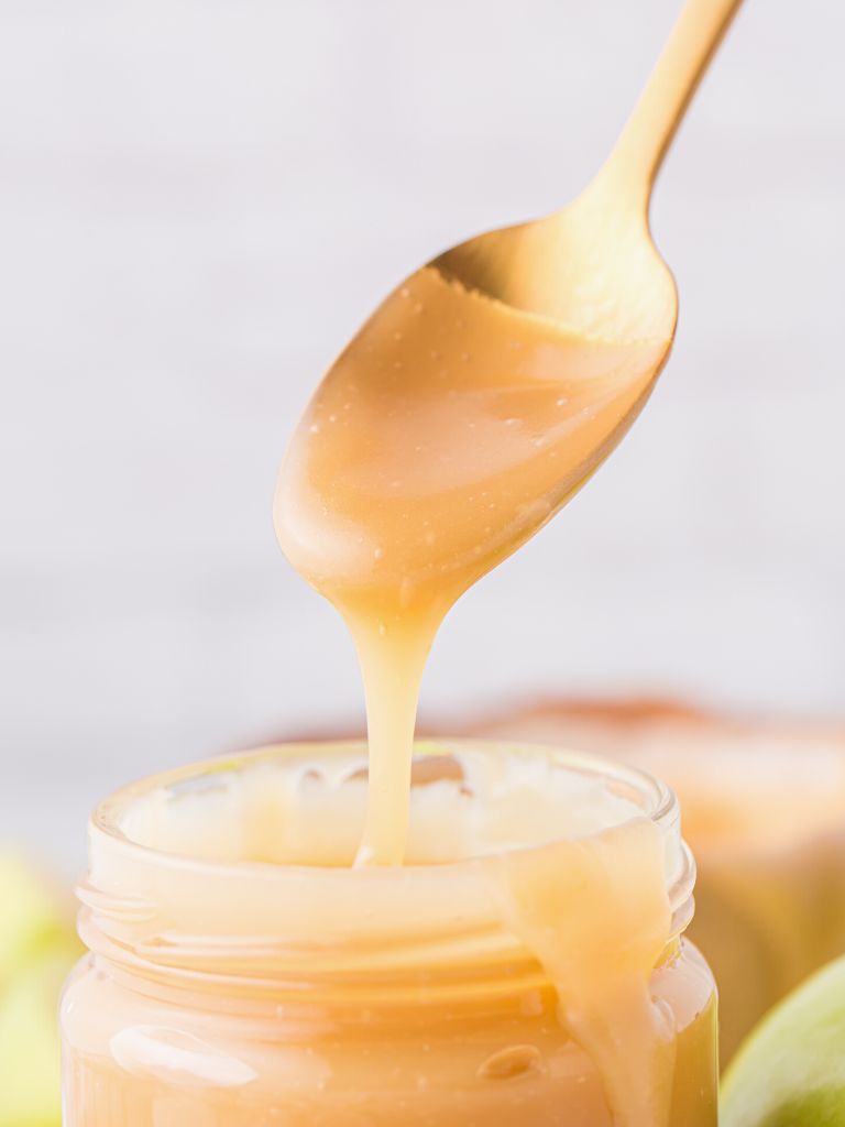 A spoonful of caramel above a jar of it, with some dripping down into the jar. 