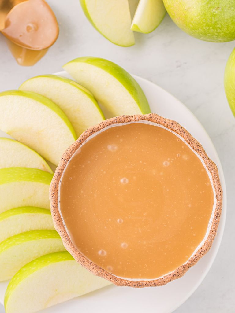 Apple slices on a white plate around a bowl of caramel dip. 