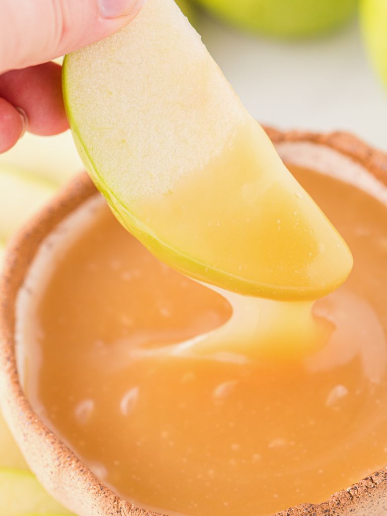 An apple slice being dipped into caramel inside a bowl. 
