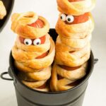 Two hot dogs made to look like mummies standing up in a black cup.