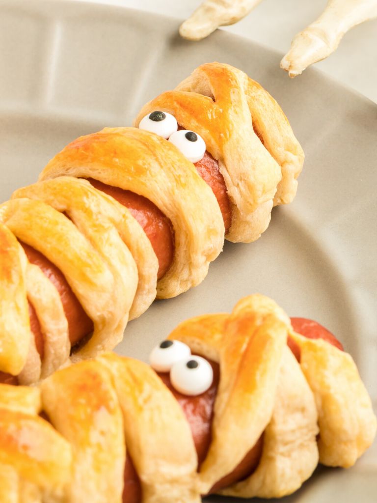 Mummy dogs on a plate with a halloween skeleton hand near it. 