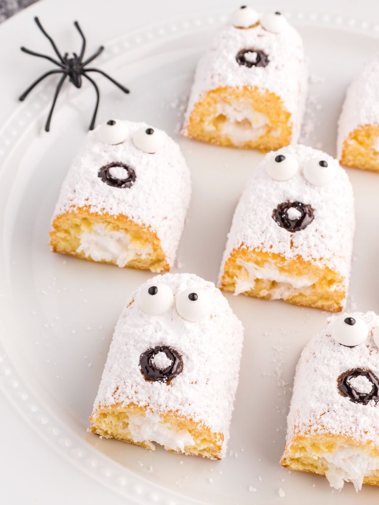 Twinkie ghosts on a white plate with a plastic fake spider next to it. 