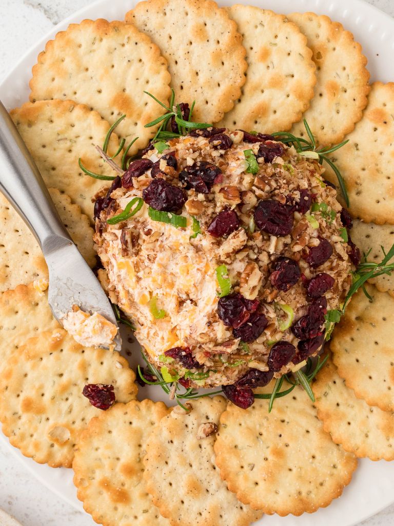 Overhead shot of a cheeseball with cheese knife next to it and lots of crackers around it. 