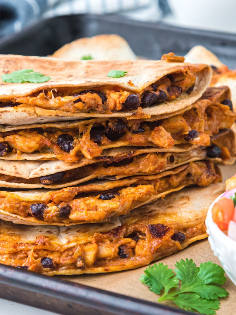 Stack of quesadillas on a sheet pan with cilantro leaves beside it. 
