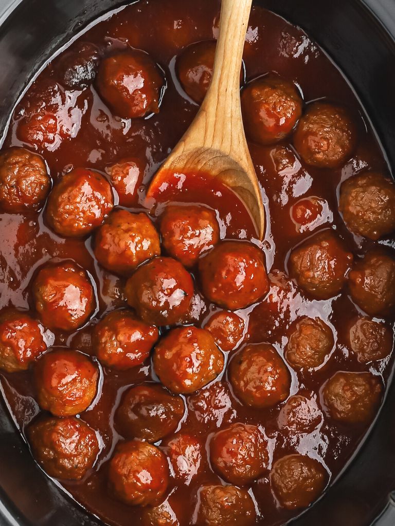 Crockpot with meatballs in a jelly sauce and wooden serving spoon. 