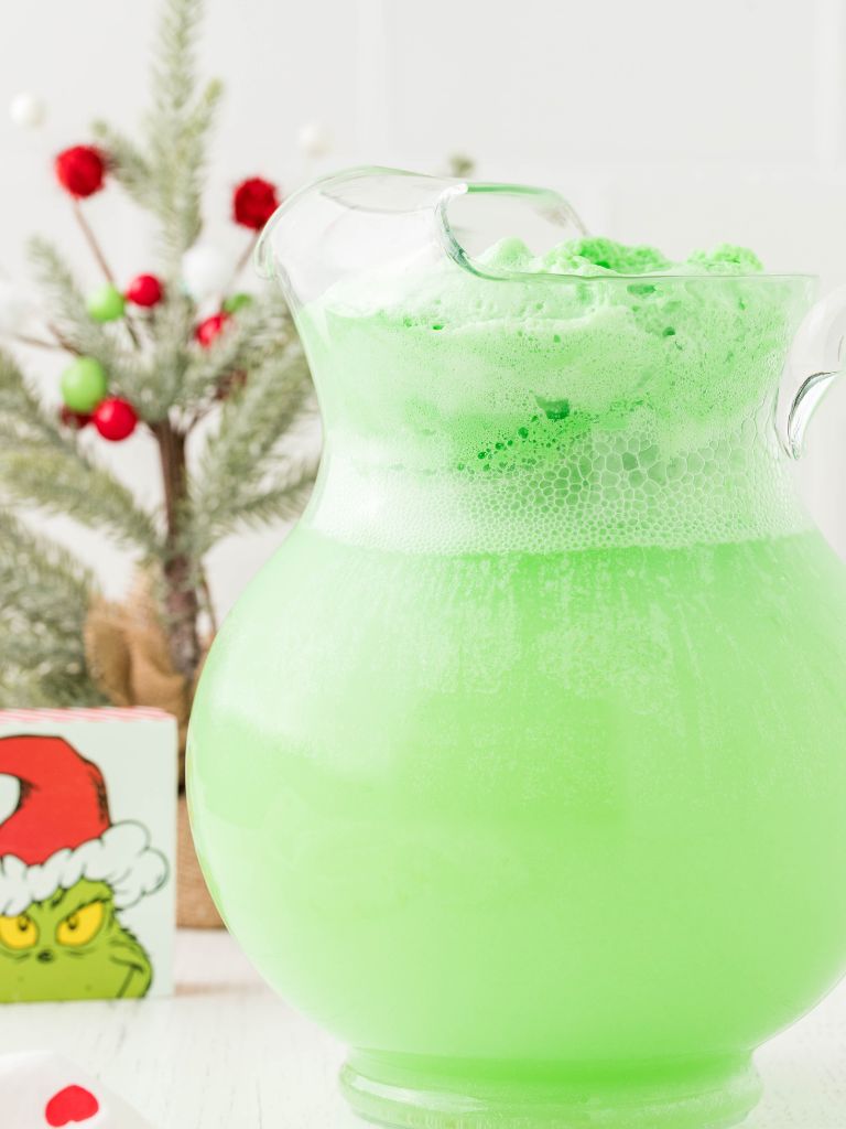 A glass pitcher of green punch with a tree and a grinch graphic in the background. 