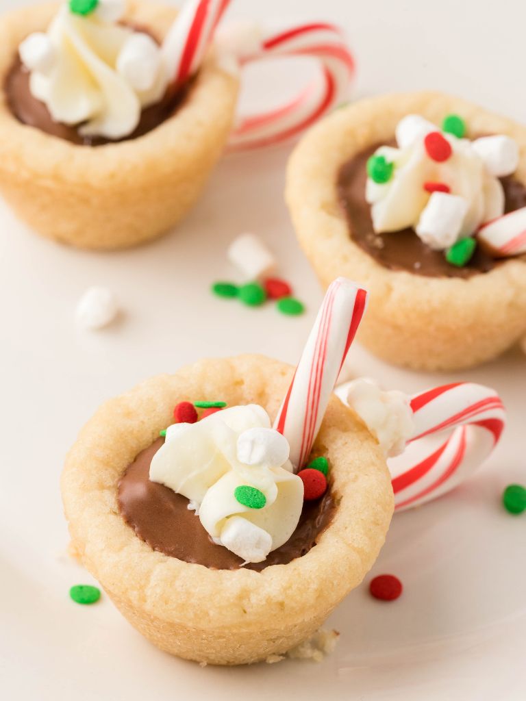 Three cookie cups sitting on a white plate made from sugar cookie dough, Reese's, and candy canes. 