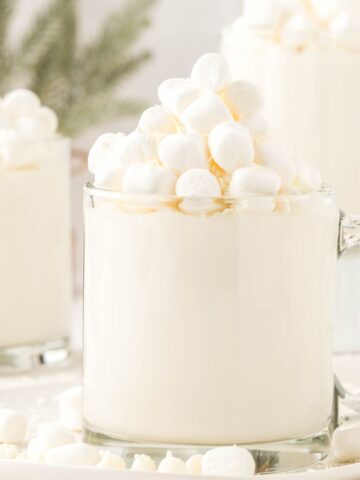A glass mug of hot chocolate that is white from white chocolate chips. Topped with mini marshmallows.