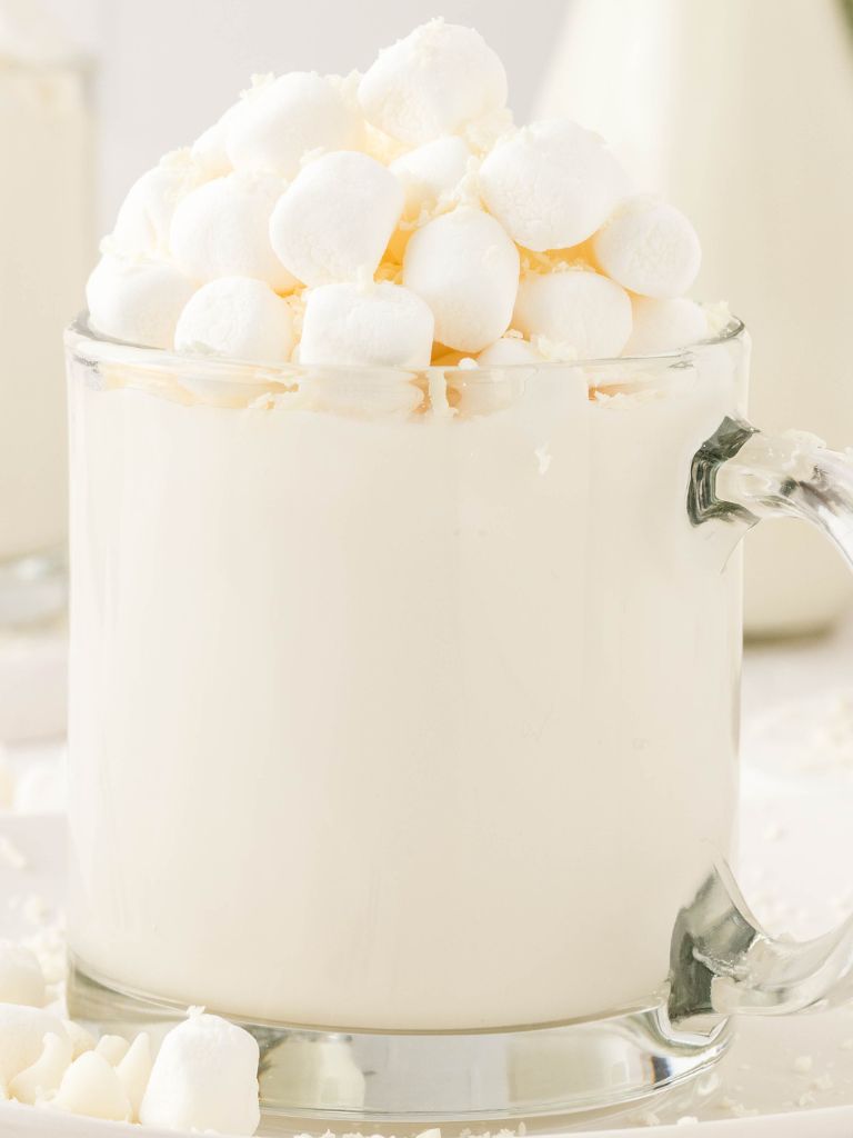 Clear glass mug of white hot cocoa topped with mini marshmallows and white chocolate curls. 